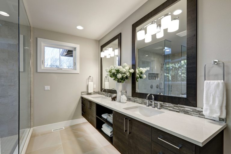 Modern coastal bathroom with mosaic tile wall, double vanity, gold fixtures, and warm lighting.