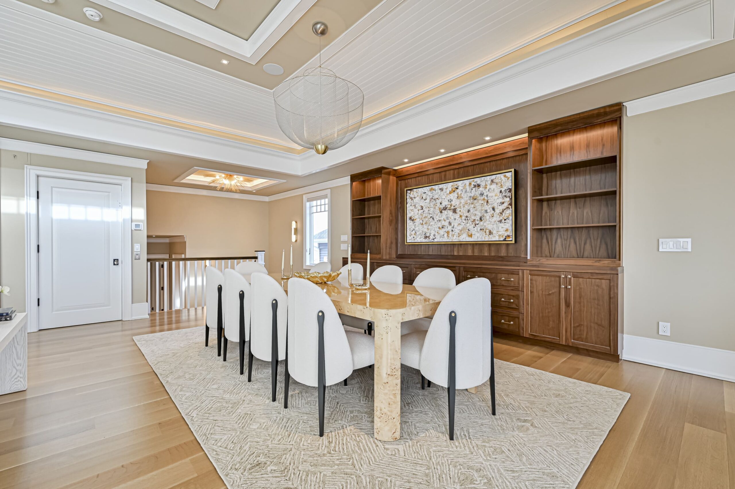 Elegant dining room with a long stone-topped table, white upholstered chairs, and a large circular metal chandelier above. In the background, a wooden wall unit with built-in shelves and artwork adds warmth to the space.