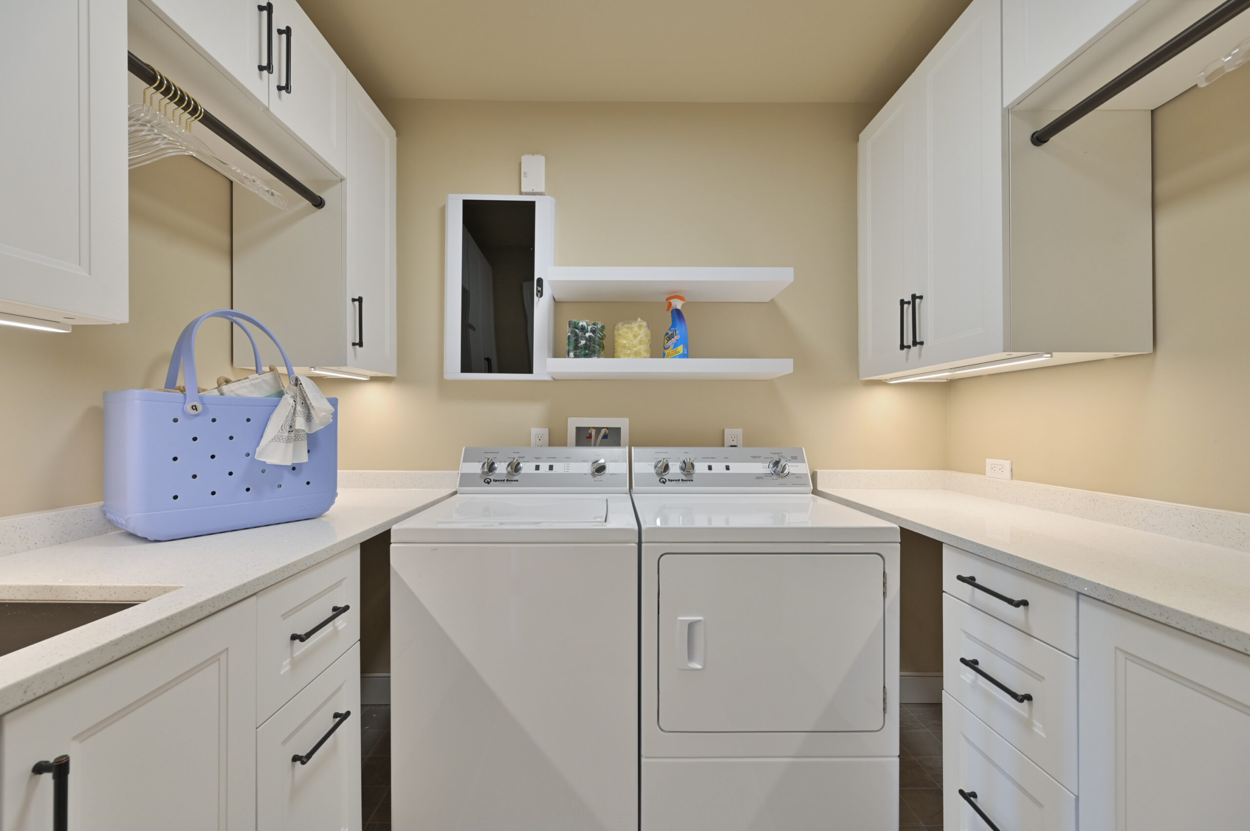 Laundry room with stacked washer and dryer under white cabinets and countertop, blue tote bag on the counter, and shelves above.
