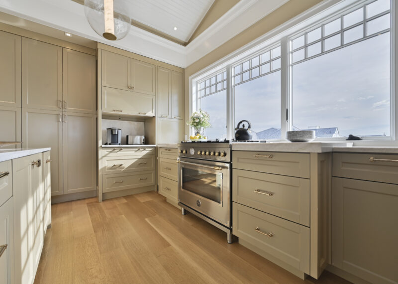 Bright modern kitchen with beige cabinets, stainless steel range, and large windows overlooking a blue sky outside.