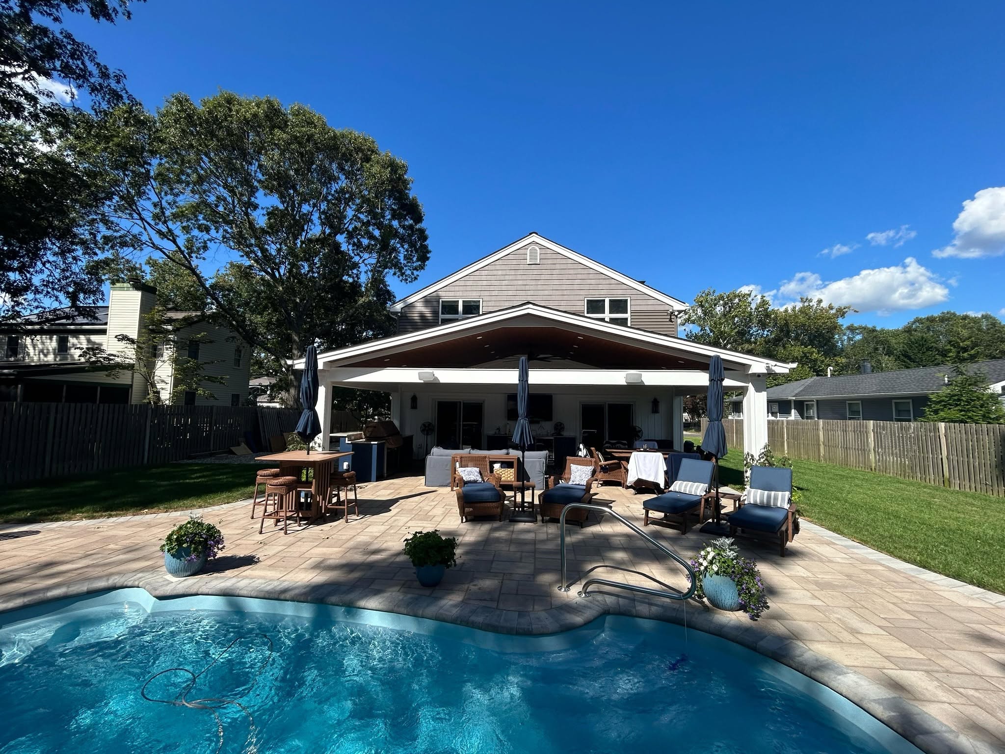 Backyard patio with poolside seating and a house in the background on a sunny day.