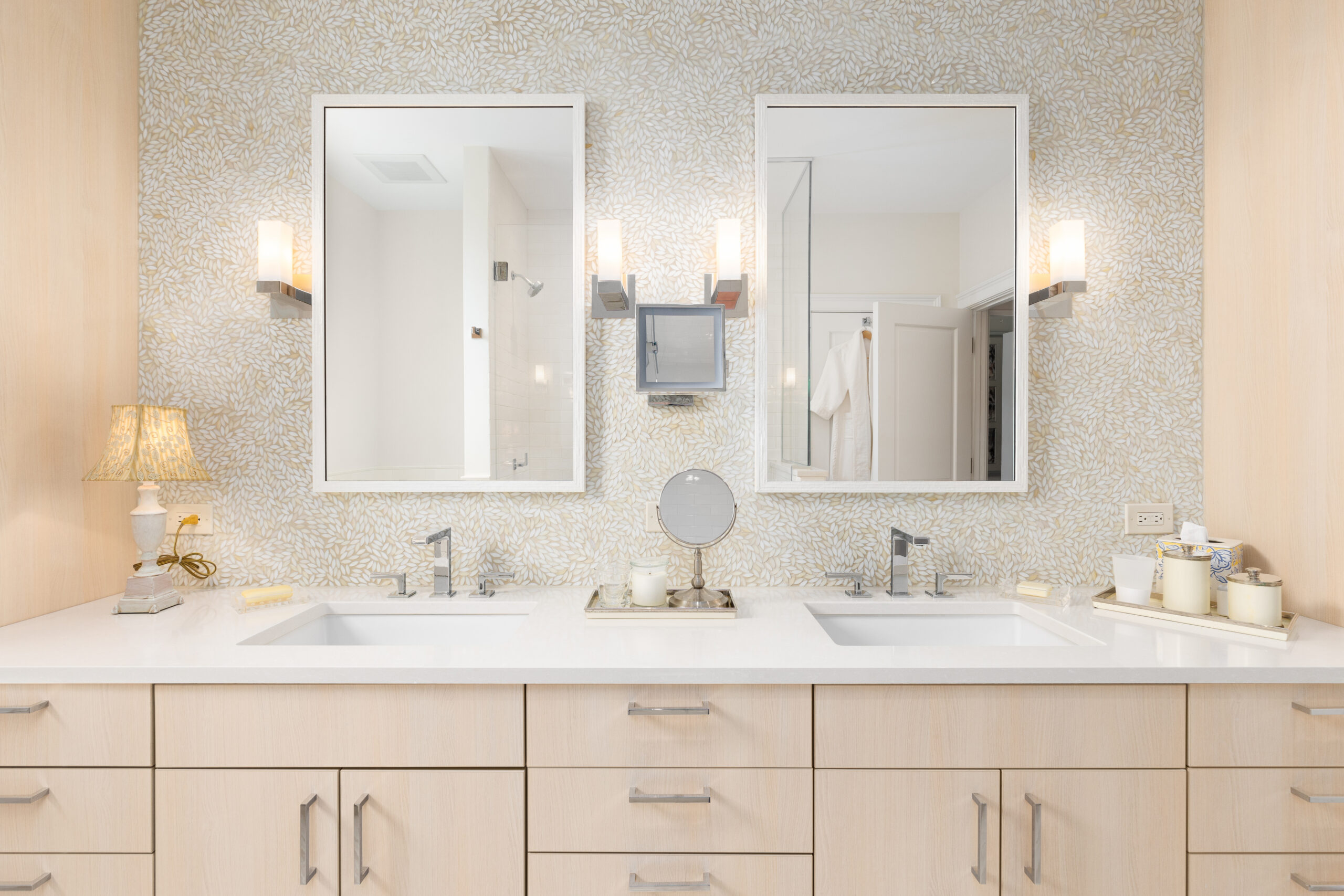 Double-sink bathroom vanity with light wood cabinets, marble countertop, and beige patterned wallpaper, flanked by three mirrors and two wall lamps in a bright, spa-like bathroom.